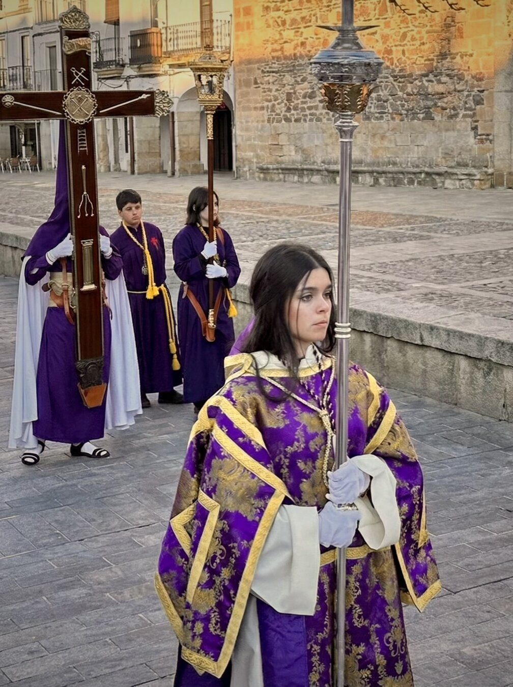 Una joven con túnica morada y dorada encabeza una procesión en Cáceres, seguida por penitentes que portan una cruz.